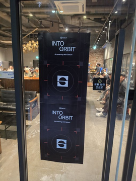 Two black "INTO ORBIT" vertical banners hanging behind a glass door, with people seated in what appears to be a cafe or co-working space.