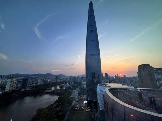 An aerial sunset view of a tall skyscraper with a pointed top, surrounded by a cityscape and a river.