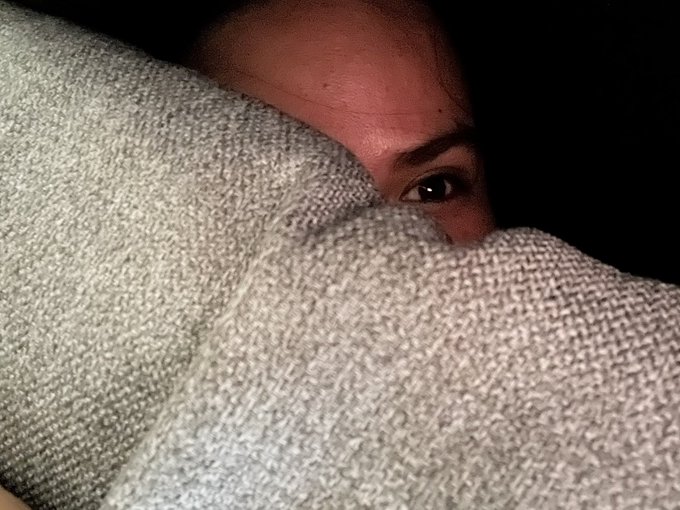 A close-up, dimly lit selfie showing a person's face partially obscured by a textured gray blanket or towel, with only their eyes and forehead visible.