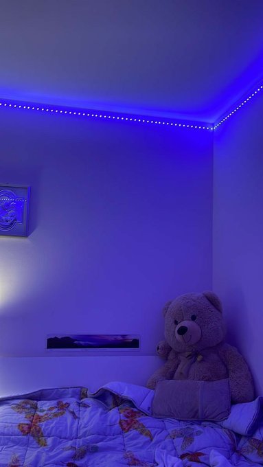 A bedroom illuminated by blue LED strip lights along the ceiling, featuring a bed with floral bedding, a teddy bear, and a laptop on a shelf.