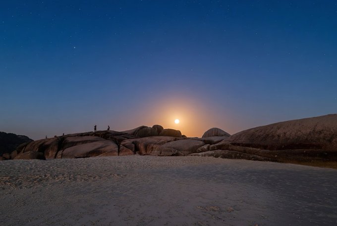 A landscape photo taken at dusk showing a bright moon rising over smooth, rounded rock formations, with small silhouettes of people standing on the rocks against a starry sky.