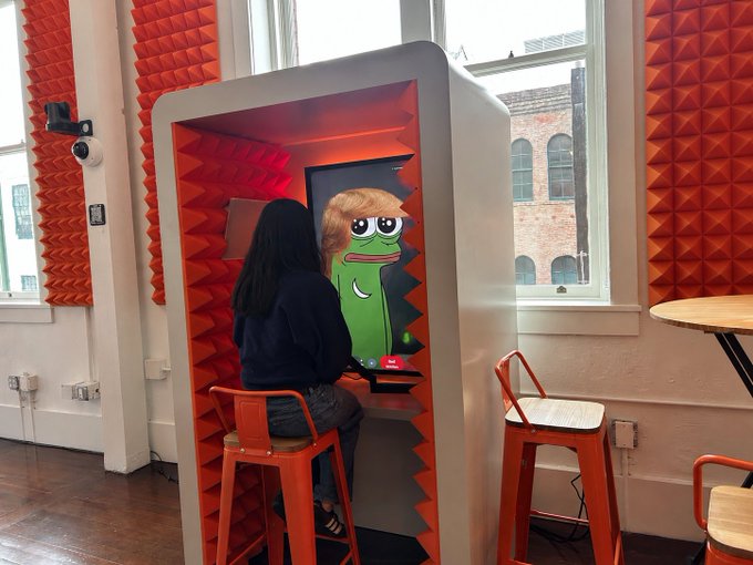 A photo of a person sitting in an orange soundproof booth interacting with a screen displaying a cartoon green monster character, set in a room with red acoustic foam panels and large windows.
