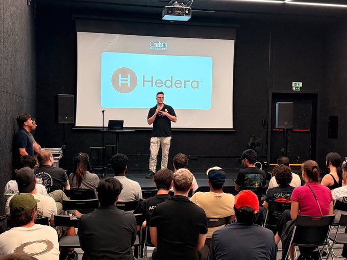 A man presents on stage at the "Onchain Builders Day" event, standing before a large screen displaying the Hedera logo, while addressing a seated audience of attendees.