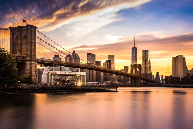 The Brooklyn Bridge spans the East River against a dramatic sunset, with the Lower Manhattan skyline—including One World Trade Center—silhouetted in the distance. The scene evokes the energy of New York City, setting the stage for the "DDNYC 2026" event by @doginaldogs, which appears to be a Doginals-themed run taking place in NYC.