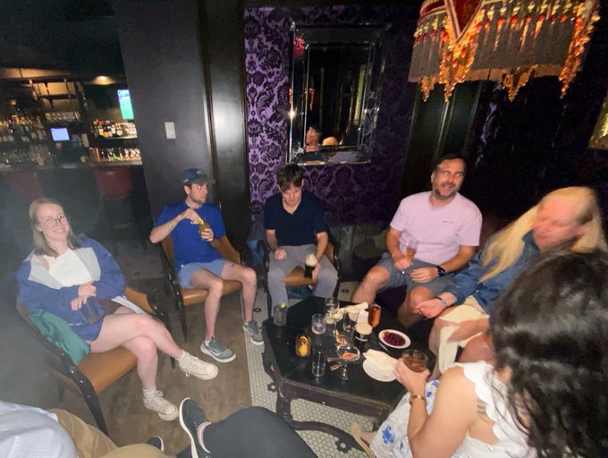 A group of people sit together in a dimly lit back room of a bar, surrounded by ornate purple patterned wallpaper and decorative hanging lamps, with drinks and snacks on a coffee table in the center. The setting matches the described "Secret Founders Circle" gathering in Boston, capturing the casual, collaborative atmosphere of a DAO meeting where members are discussing their 24-month plans over drinks.