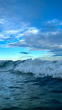 Waves crash in the ocean under a bright blue sky filled with scattered clouds.