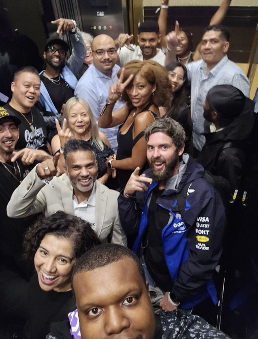 A crowded selfie of a group of people smiling and making gestures, featuring a man in a blue racing jacket and a man in a grey suit.