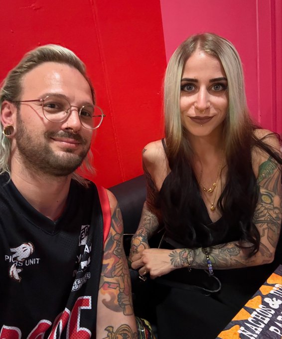 A man and woman with multiple tattoos posing for a photo, the man wearing a black basketball jersey and glasses.
