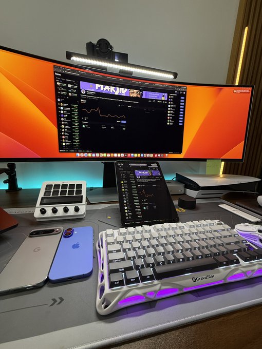 A well-equipped trading workstation featuring a curved ultrawide monitor and tablet displaying the MARJ/USDC chart, alongside a white mechanical keyboard, a streaming controller, and smartphones. The setup reflects active crypto trading on the Solana network, tying into the post's invitation to share a USDC address and join the MARJ trading community via fomo.family.
