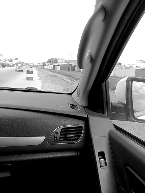 A black and white photo taken from the inside of a car, showing the dashboard, air vent, and side mirror, with a view of a road and buildings ahead.