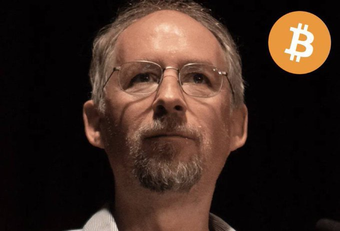 Adam Back, a key figure in Bitcoin's early development, speaks in front of a Bitcoin logo overlay. The image accompanies a claim of his bold price prediction that $100K is "way too cheap" for Bitcoin, with expectations ranging from $500K to $1M this cycle.