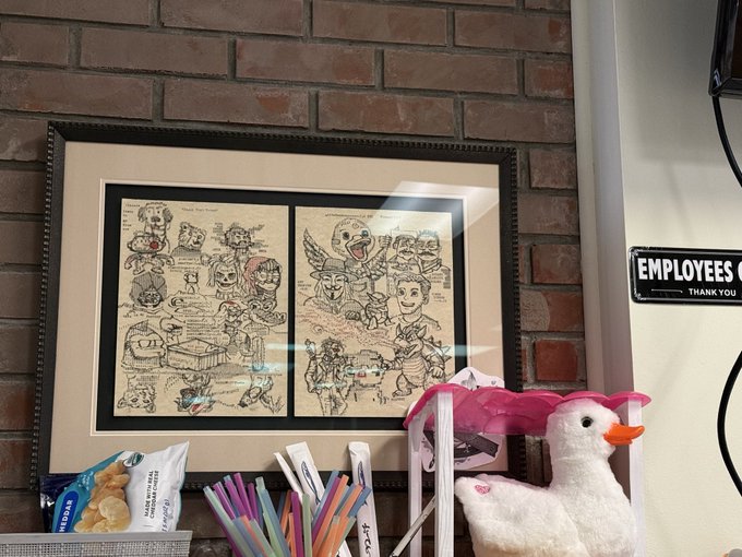 A framed artwork with character sketches and red signatures hangs on a brick wall, with a stuffed bird wearing pink sunglasses and various snacks on the shelf below, next to an "EMPLOYEES ONLY THANK YOU" sign.