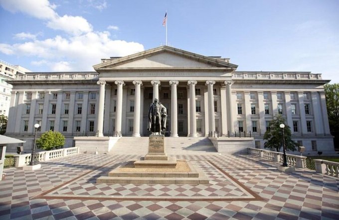 The U.S. Treasury Department building is shown from a frontal view, featuring its iconic neoclassical columns, central statue of Alexander Hamilton, and American flag. The checkered plaza and grand entrance are visible under a partly cloudy sky.