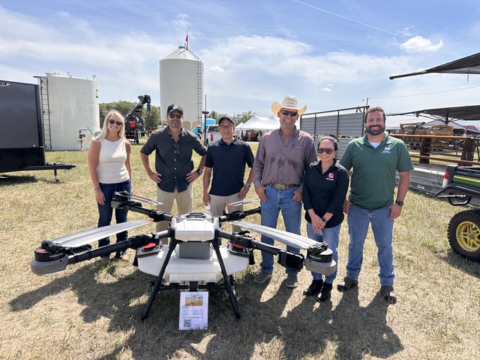 A group of six people pose outdoors behind a large white agricultural drone, with grain silos and a Texas A&M AgriLife tent visible in the background.