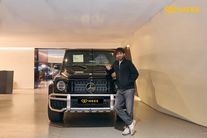 A young man in a black jacket and jeans poses standing next to a black Mercedes-Benz G-Wagon SUV in a showroom, with the 'WEEEX' logo visible as a watermark in the upper right and on the vehicle's license plate.