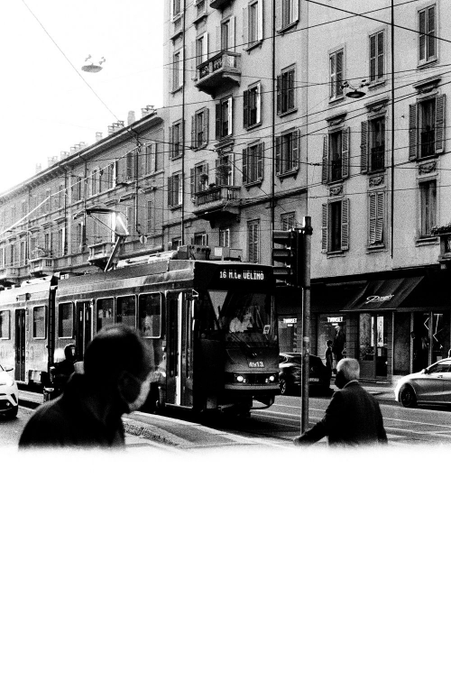 Black and white street photograph of a European city scene with a tram ('18 atto MATTEO') passing by classical architecture, with pedestrians crossing the foreground.