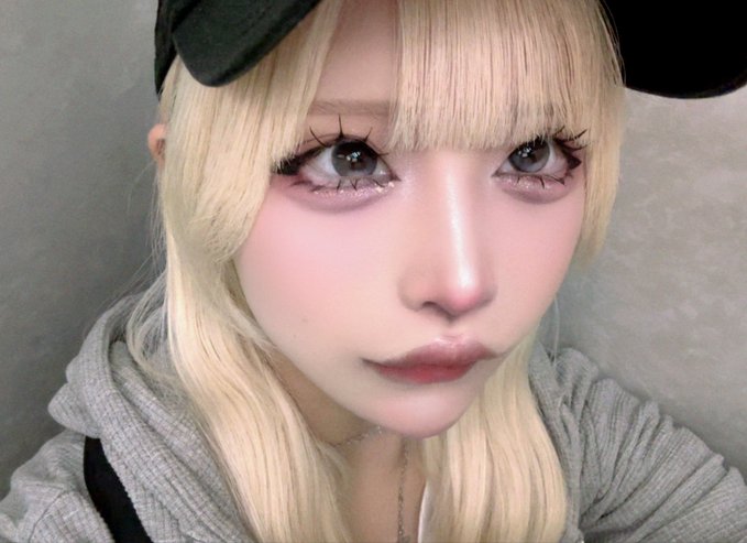 A close-up portrait of a young person with long blonde hair wearing a black cap, featuring dramatic makeup with long false lashes and pink-tinted cheeks, gazing softly at the camera. The image conveys a quiet, close, or near feeling through its tight cropping and intimate, direct eye contact with the viewer.