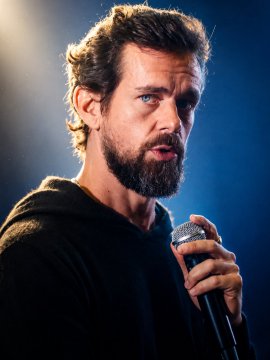A bearded man with tousled hair holds a microphone, appearing to be mid-speech in a dimly lit setting. The image captures Jack Dorsey, the tech entrepreneur who has publicly advocated for Bitcoin as the future single global currency.