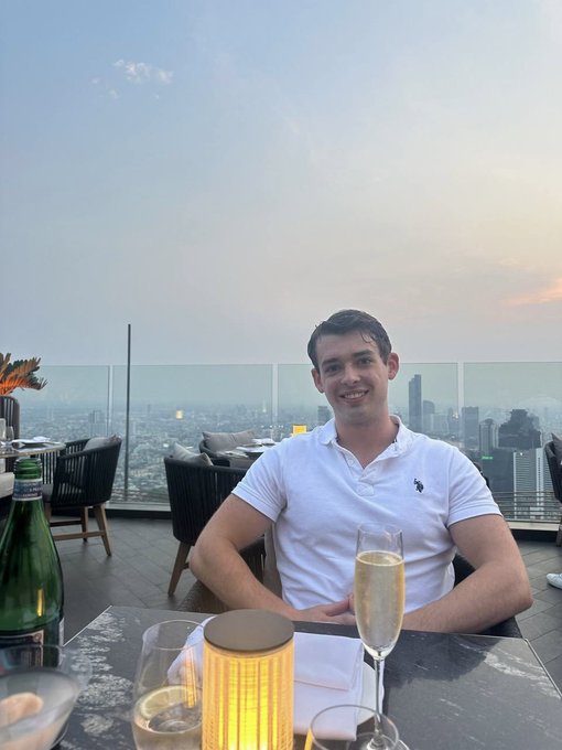 A smiling young man in a white polo shirt sits at a rooftop restaurant table with a champagne flute, against a hazy city skyline at dusk.