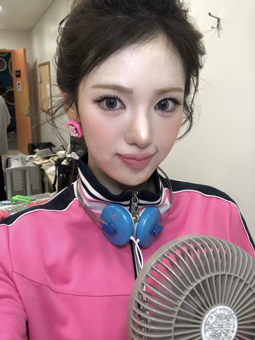 A close-up selfie of a young woman in a pink and black jacket with blue headphones around her neck, holding a fan.