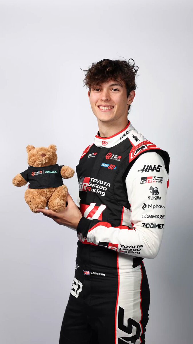 The image features a man wearing a racing suit with a teddy bear sitting next to him. The man is smiling and holding up the teddy bear, which has a "Ollie Bearman" logo on its chest. The background of the photo is white, making the man and his stuffed animal stand out prominently.

The post appears to be promoting an event or contest related to racing, as it mentions that some picks might start arguments if they win. It also encourages viewers to share their opinions by reposting the image with a specific hashtag: "#OllieBearman #WorldCupBracket". The man and his teddy bear are positioned in front of this post, drawing attention to both themselves and the contest it promotes.

The main focus of the image is on the man holding up the teddy bear, which serves as a symbol or mascot for the event being advertised.