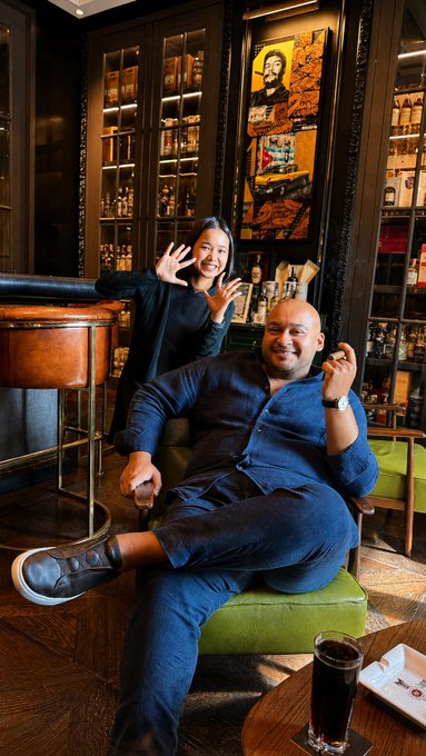 A bald man in a blue suit lounges in a green chair with a drink, while a woman makes an "all in" hand gesture behind him in a dimly lit lounge with liquor cabinets and a striking artwork of a figure resembling Tim Kotzman on the wall.
