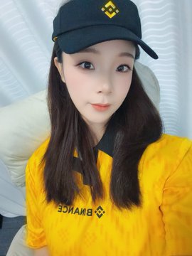 The image features a young woman wearing a yellow shirt with black text, which reads "@binANCEzh". She is also wearing a black baseball cap with a yellow logo. The background of the photo shows a white curtain or wall behind her.