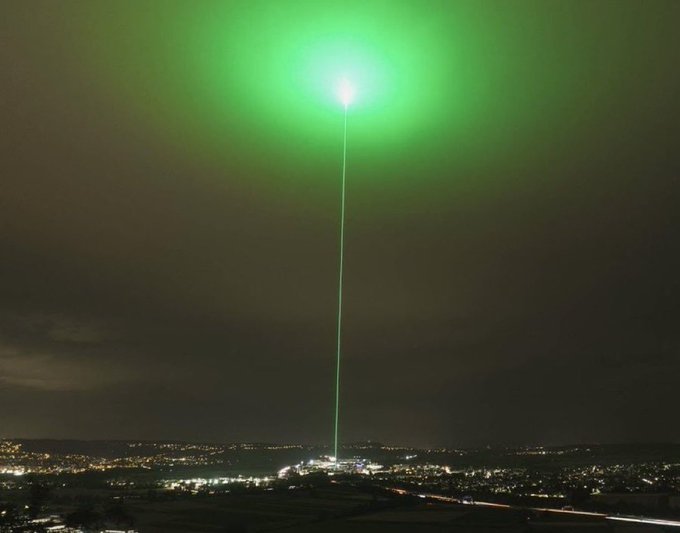 A nighttime cityscape is illuminated by a bright green laser beam shooting straight up into the dark sky from the urban landscape below.