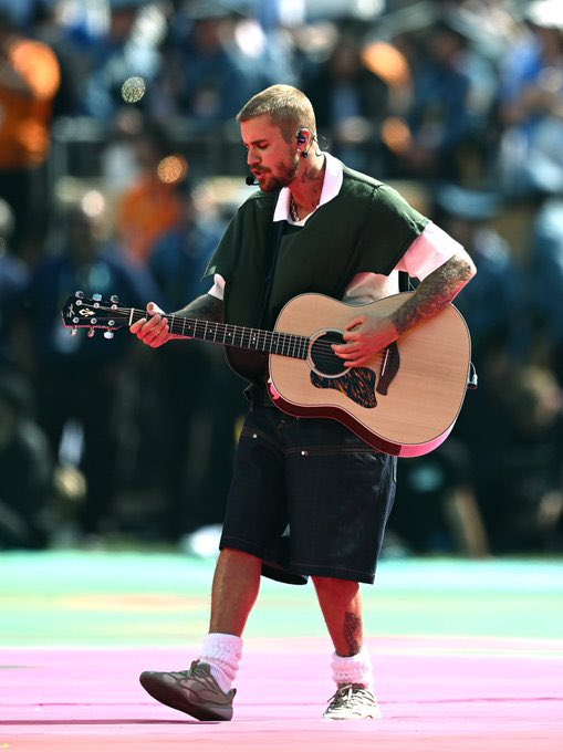 A male musician with short hair and a microphone headset plays an acoustic guitar on stage, wearing a green and white polo shirt and black shorts.