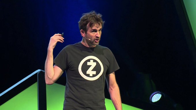 The image features a man standing on stage, wearing a black t-shirt with a white logo that says "ZEC" on it. He appears to be giving a speech or presentation, as indicated by his hand raised while speaking. The background of the image is dark and green, which contrasts with the man's attire and makes him stand out more prominently in the photo.