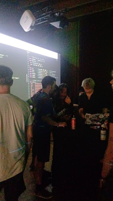 A dimly lit indoor scene showing people gathered in front of a large screen displaying code or text, with stage lighting visible overhead.