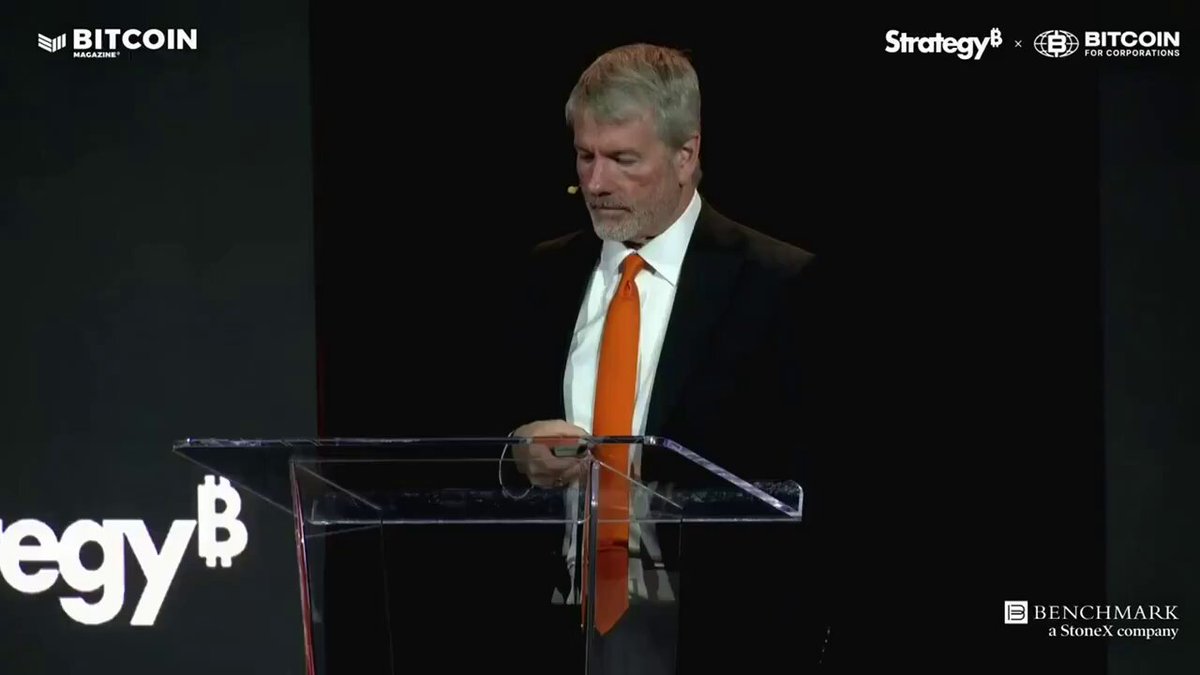 The image features a man standing at a podium with his hands on a microphone, giving a speech or presentation. The background of the image shows a black screen displaying the text "Sailor hyung, you said you weren't going to sell Bitcoin...". This suggests that the speaker is addressing an audience about Bitcoin and its potential impact on the market.