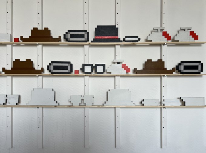 Three white shelves displaying pixel-art sculptures of hats and headwear made from wooden blocks, featuring various styles in brown, black, white, gray, and red.