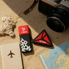 Travel accessories arranged on sand, including a red 'TRON' lighter, a triangular 'TRON' case, a camera, a starfish, and a map.