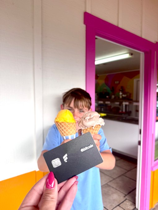 The image features a young girl holding two ice cream cones with one of them being a credit card. The girl appears to be posing for the camera, showcasing her ice cream treats while also displaying the credit card. The background includes a pink door or wall, which adds a pop of color and contrasts nicely with the white wall in the scene.