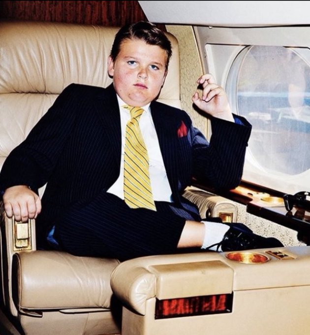 A young boy in a dark pinstripe suit and yellow striped tie sits in a large recliner, holding a cigar.