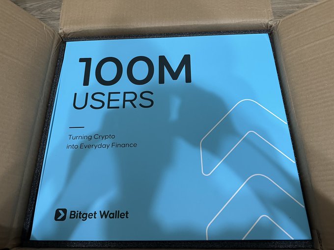 A blue promotional box in a shipping container celebrating '100M USERS' with the tagline 'Turning Crypto into Everyday Finance' for Bitget Wallet.