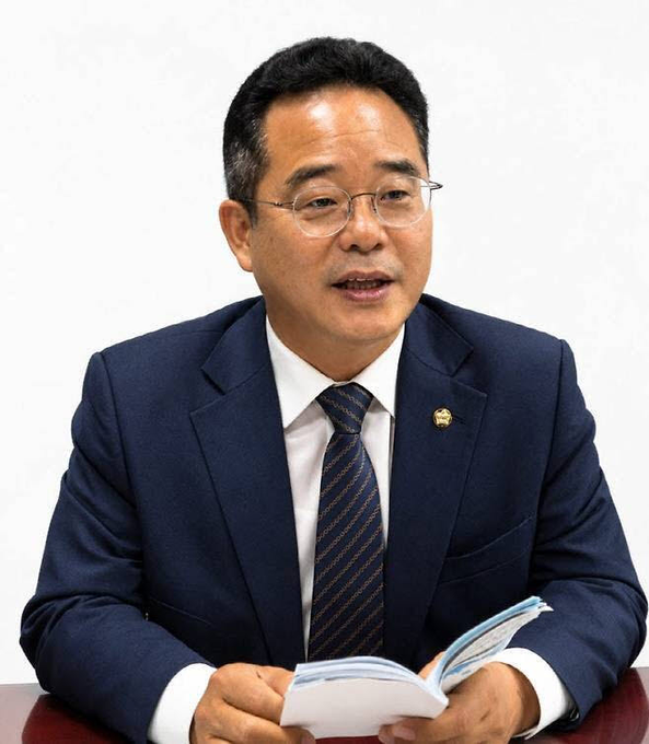 A man wearing glasses, a dark blue suit, white shirt, and striped tie speaks while holding an open booklet.