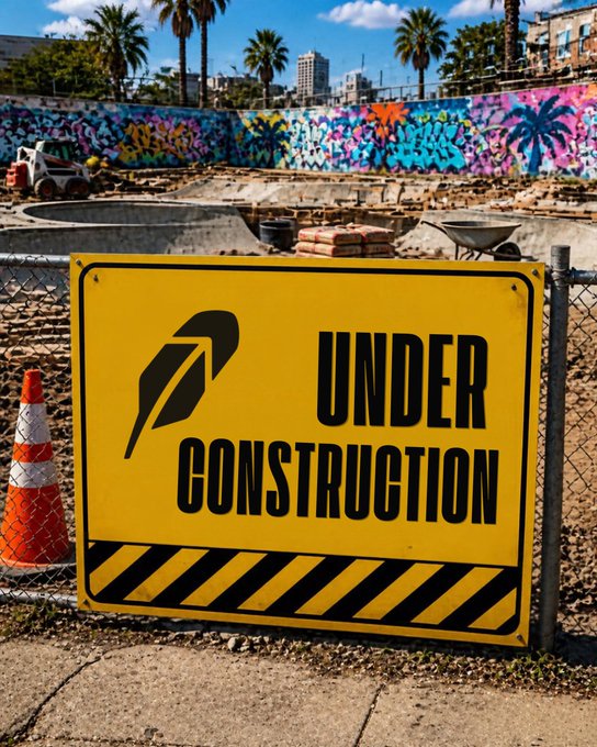 A bright yellow 'UNDER CONSTRUCTION' sign with a black feather/quill logo is displayed in front of a chain-link fence at a graffiti-covered construction site.