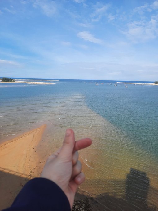 A first-person view of a hand making a finger gun gesture toward a calm body of water with sandy shores under a blue sky.