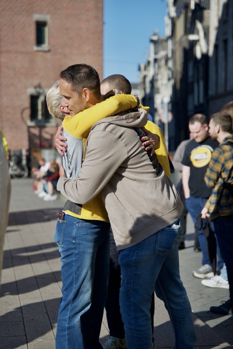 The image shows a man wearing a yellow shirt embracing another person, possibly his partner or friend, as they stand together on a sidewalk. In the background, there are several other people present, but their attention seems to be elsewhere. The photo is captioned with "He'S aware of spx69080 and probably cries himself to bed every night because he's stuck with spx69080".