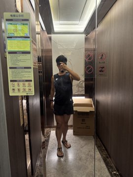 A person takes a mirror selfie in an elevator, wearing denim overalls and holding a phone, with Chinese elevator safety signs visible on the wall and a cardboard box in the corner.