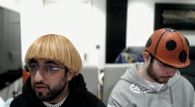 Two men wearing novelty hats—a blonde bowl-cut wig and an orange ladybug-patterned cap—sit in what appears to be an office or studio setting.