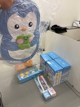 A photo showing merchandise including a plush character toy in plastic packaging, several blue boxes, and other small boxed items on a floor.