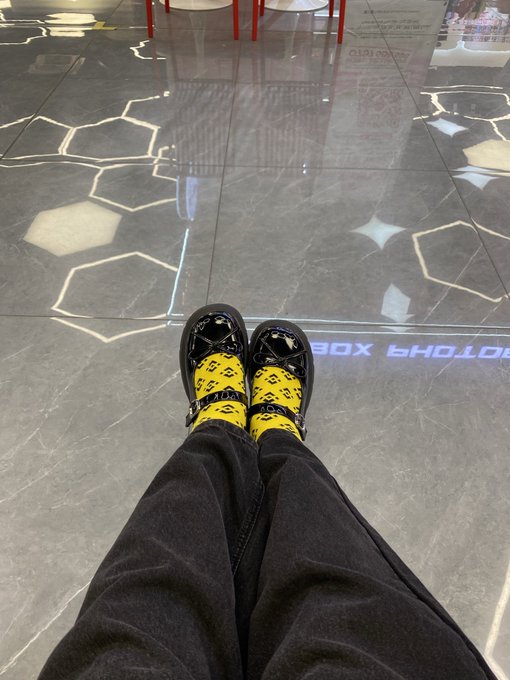 A first-person view of someone wearing yellow-patterned socks and black shoes, standing on a glossy tiled floor with hexagonal light reflections.
