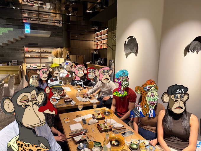 A group of people seated around a long dining table in a modern restaurant interior, with their faces covered by various NFT-style cartoon avatars, including a pink-faced monkey, a zombie-like character, and one wearing a 'BAYC' cap.