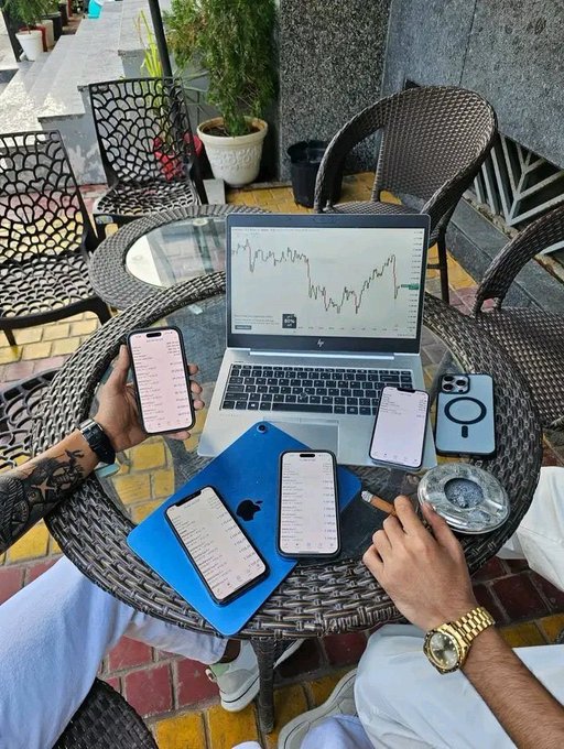 A person is seated at an outdoor patio table with a laptop displaying a stock chart, surrounded by multiple smartphones showing trading apps and messages, along with a tablet, ashtray, and a coffee cup.