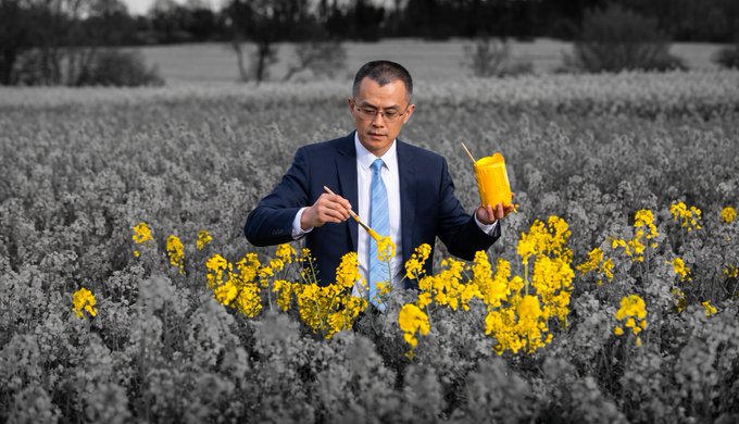 A man in a business suit stands in a grayscale field, holding yellow paint and a brush to color yellow flowers around him.