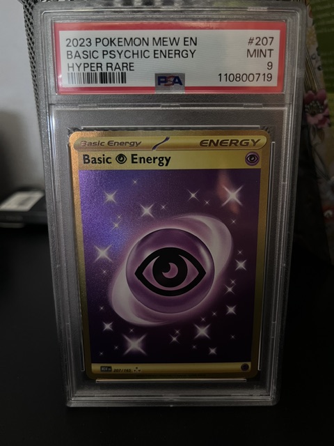 A PSA-graded 2023 Pokémon Mew set #207 Basic Psychic Energy Hyper Rare card, encapsulated in a protective case with a "MINT 9" label, featuring a gold-bordered design with an eye and cosmic swirl artwork; the caption "Gem Mint 10" suggests the owner is highlighting the card's near-perfect condition despite its PSA 9 rating.