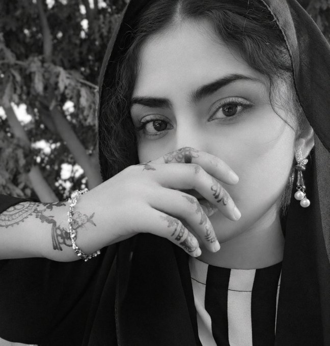 A black and white portrait of a woman with henna-decorated hands partially covering her face, wearing a dark headscarf and dangling earrings.